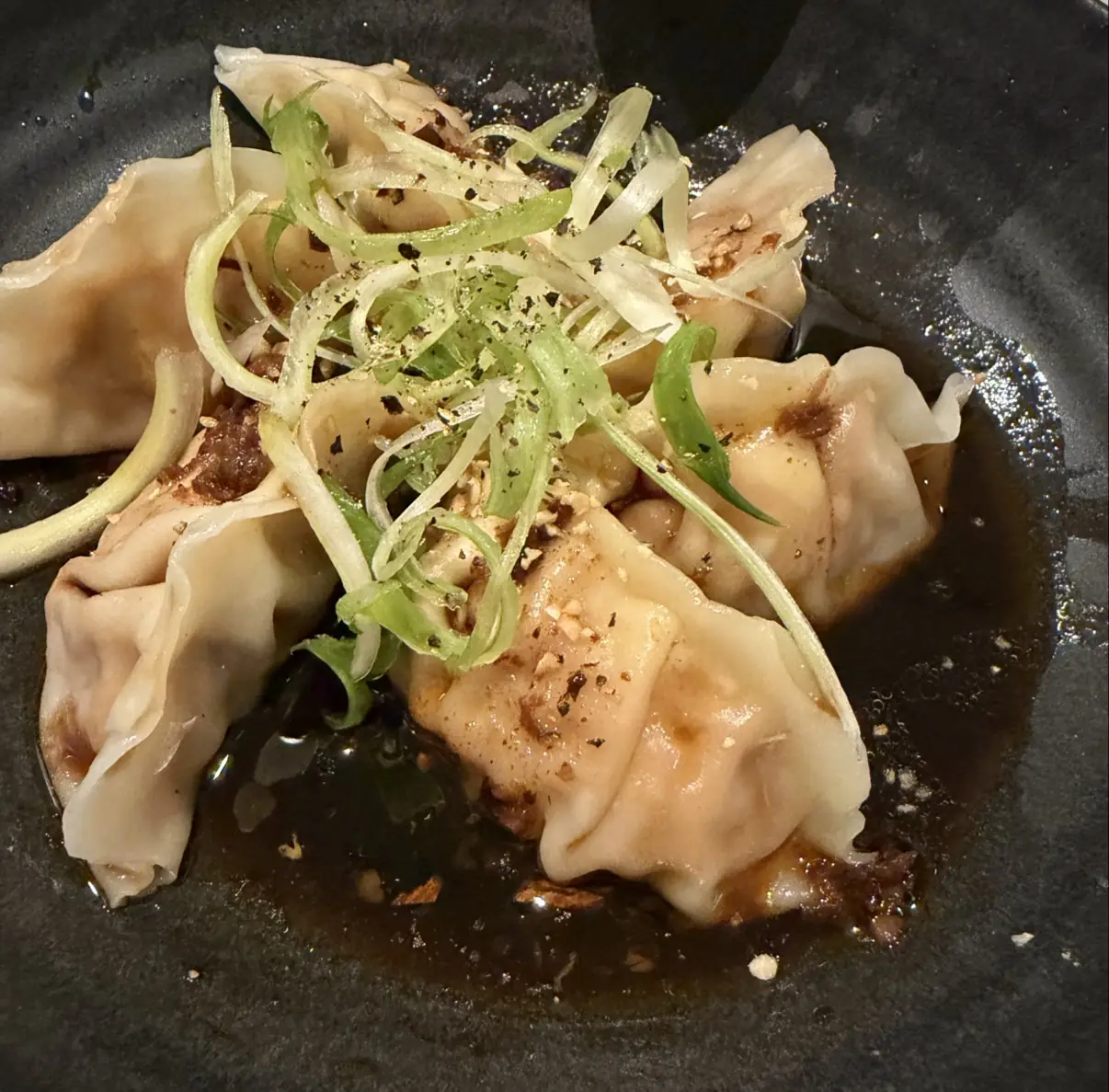 Dumplings in dark sauce topped with julienned scallion and crushed nuts on a black plate