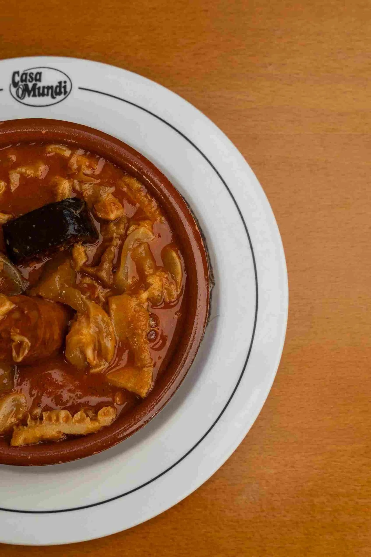 A clay dish of tripe stew with a black sausage on a white plate on a wooden table