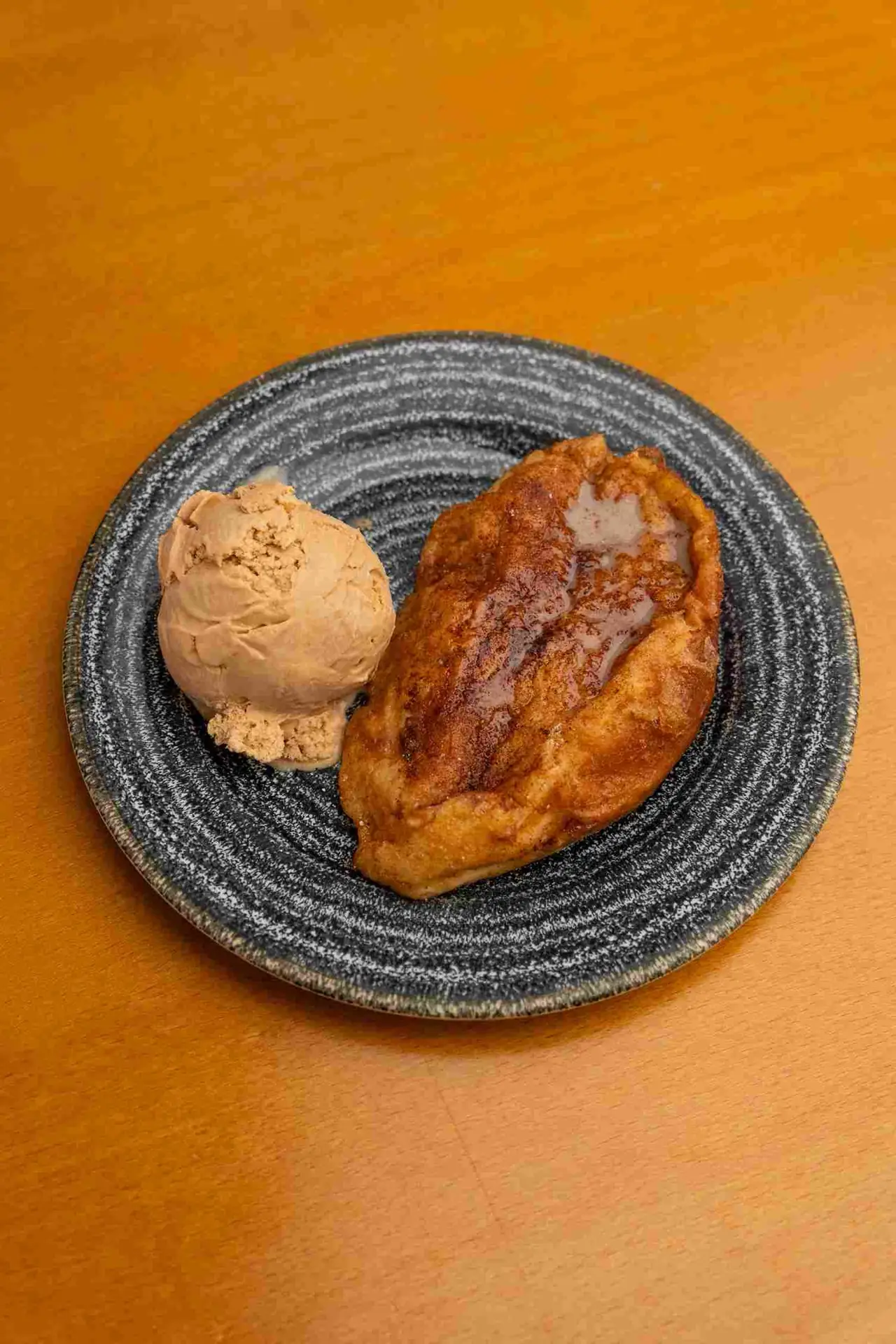 Fried torrija dusted with cinnamon beside a scoop of caramel-colored ice cream on a black speckled ceramic plate on a warm wooden table