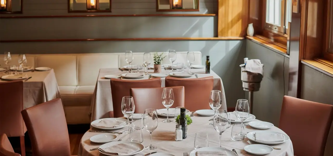 Dining room with round tables set with wine glasses and white plates, tan leather chairs and a cream banquette