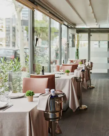 Window-side dining row with tan chairs, small potted plants and wine coolers on stands beside floor-to-ceiling glass