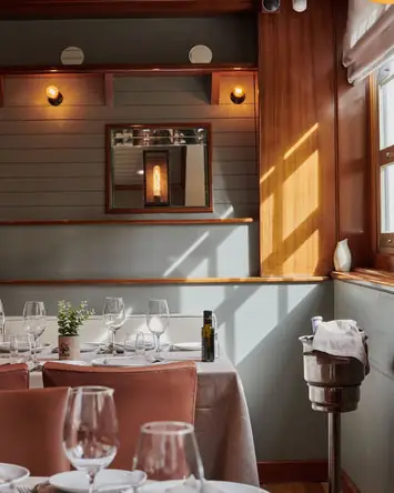 Dining-room corner with wall lamps, a framed mirror on grey panelling and set tables in sunlight