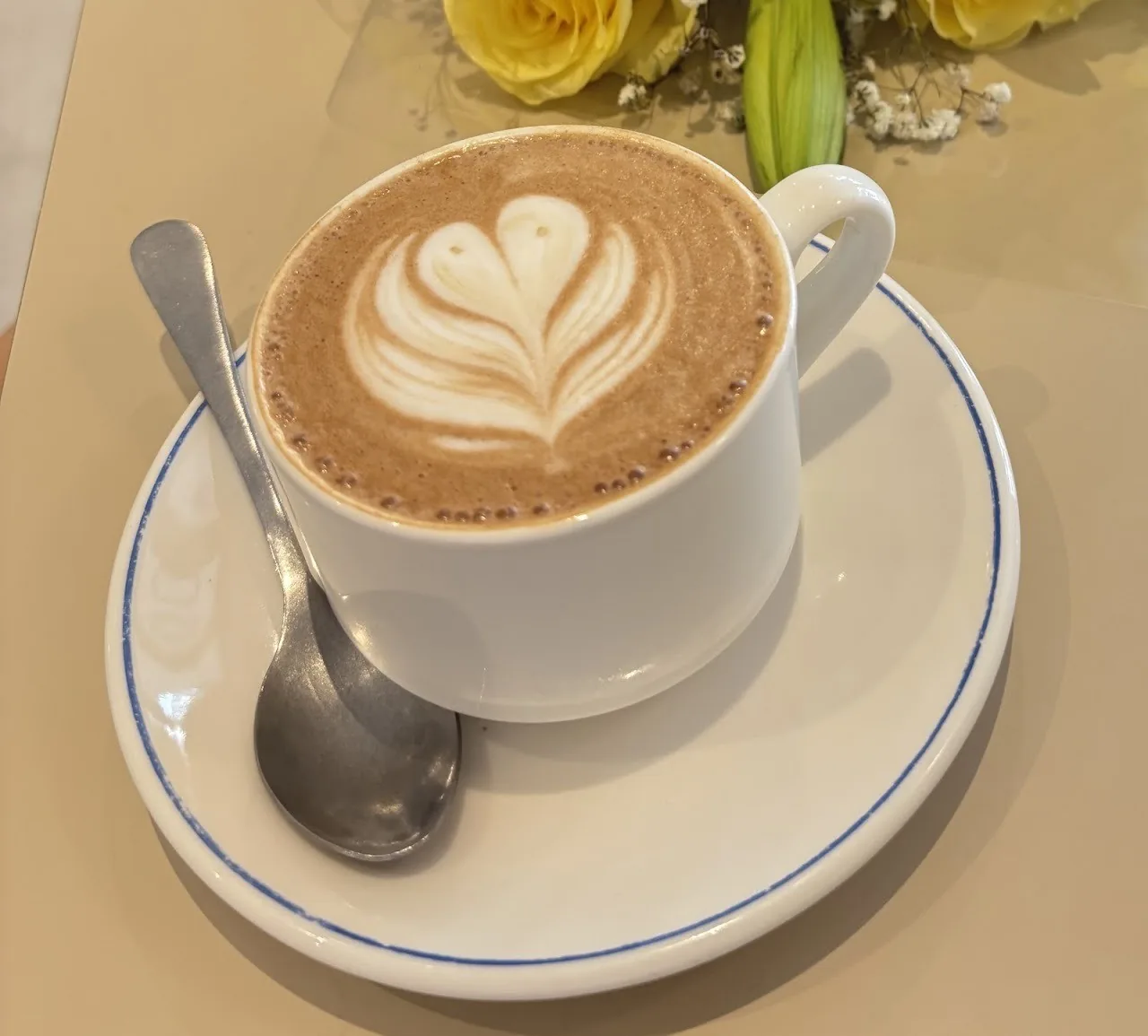 A cappuccino with a leaf pattern in a white cup with a blue rim on a saucer