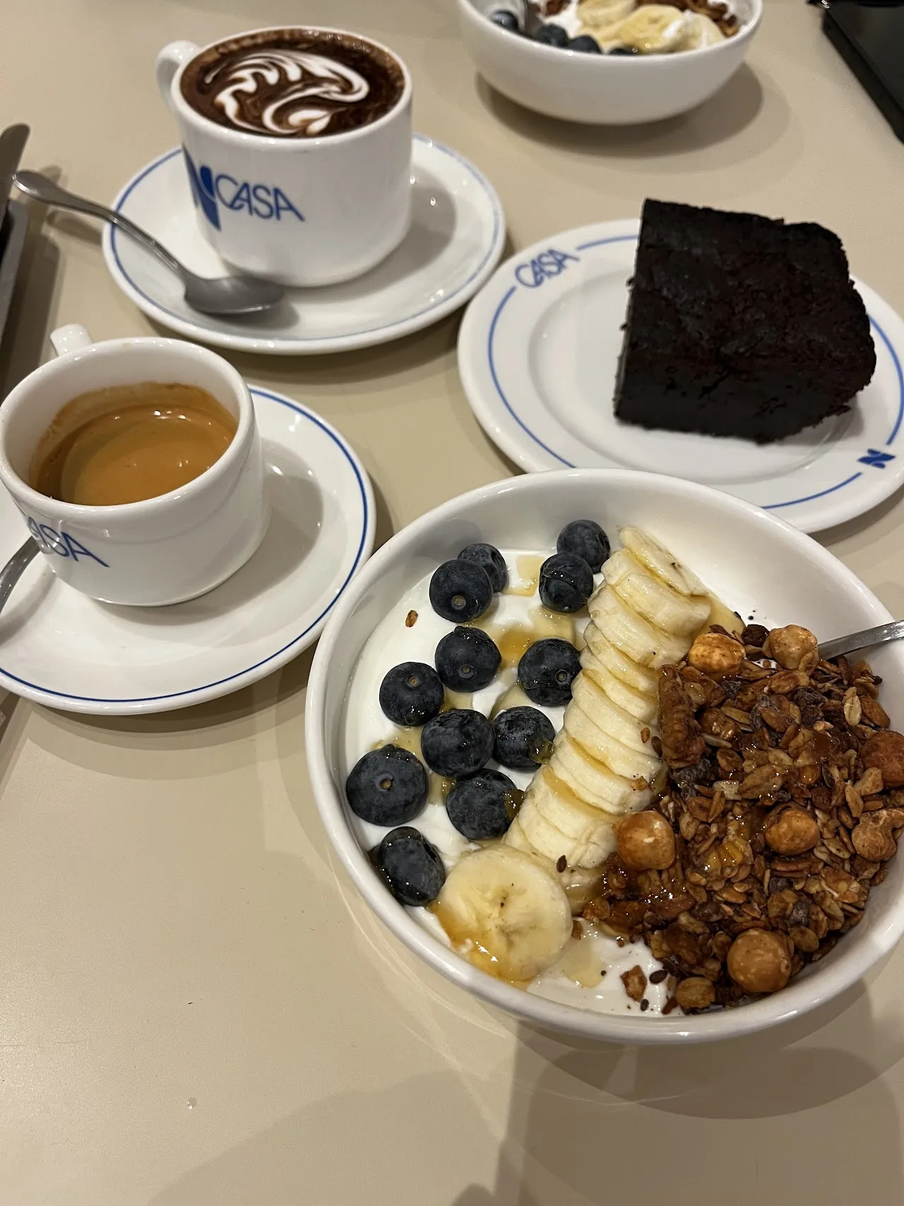A cappuccino, a slice of chocolate cake, and a bowl of yogurt with fruit and granola on a table