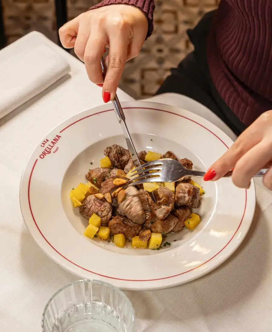 Hands using a knife and fork to cut diced sauteed meat with potato cubes and mushroom on a white plate branded Casa Orellana