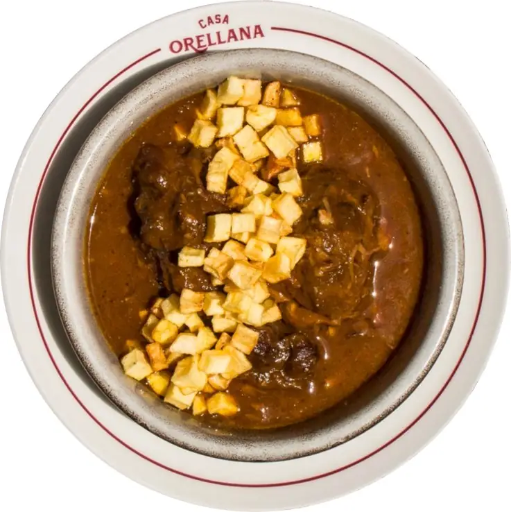Overhead shot of a stewed meat dish in dark sauce topped with diced fried potatoes, on a white plate branded Casa Orellana