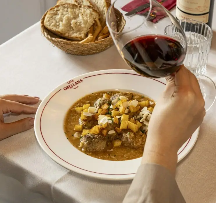 Meatball stew with diced potatoes and almonds on a Casa Orellana branded plate, bread basket and a hand pouring red wine into a glass on the table