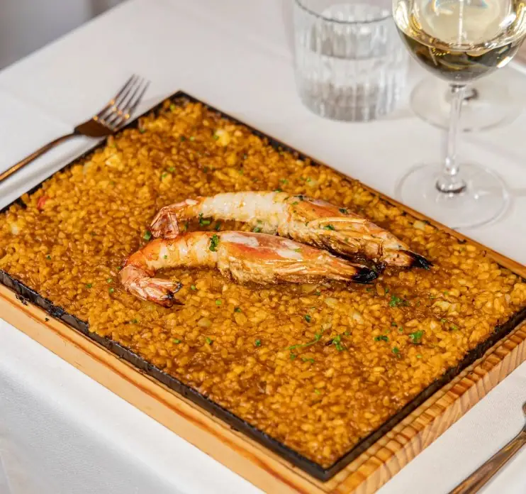 Rectangular tray of seafood rice topped with two grilled whole prawns, with a glass of white wine on a white tablecloth