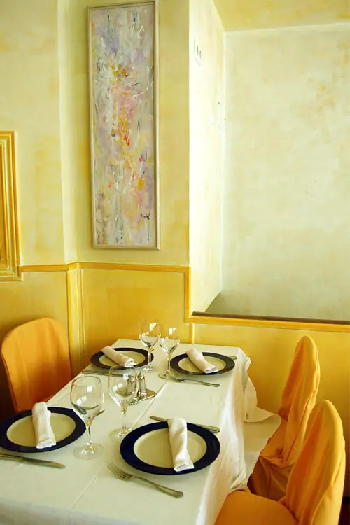Yellow-walled dining corner with a set table dressed in white linen with navy-rimmed plates, orange chair covers and a tall abstract painting