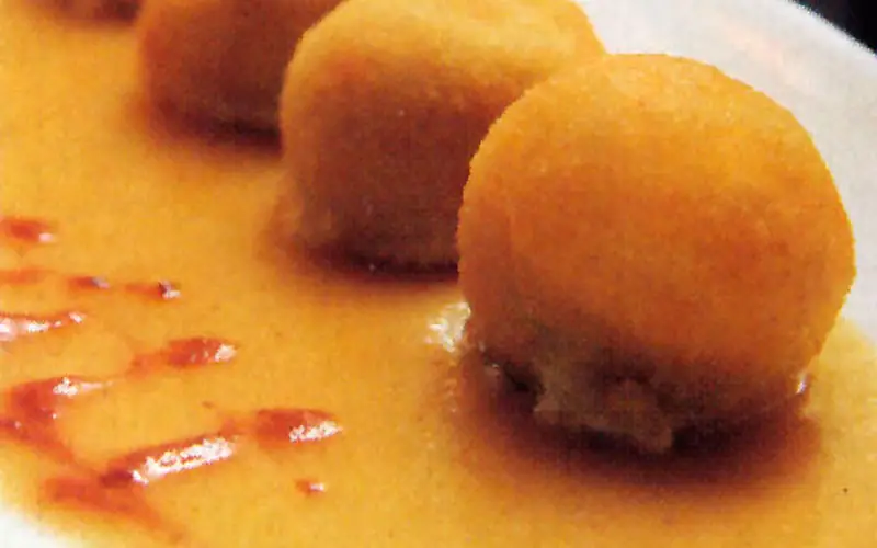 Row of golden fried croquette-style balls sitting in a creamy orange sauce with a swirl of red sauce