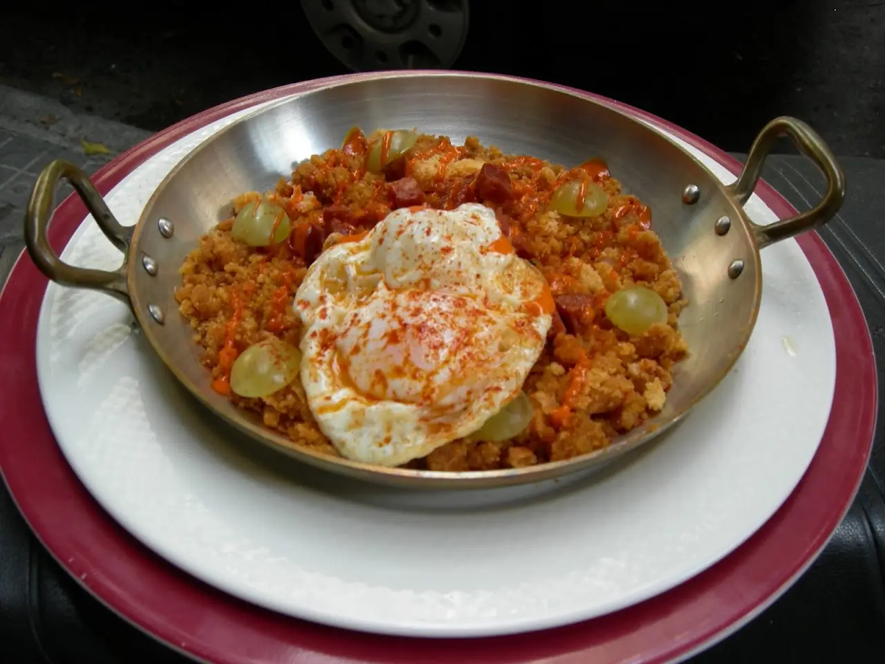 A fried egg over a pan of migas (fried breadcrumbs) with grapes, in a copper serving pan