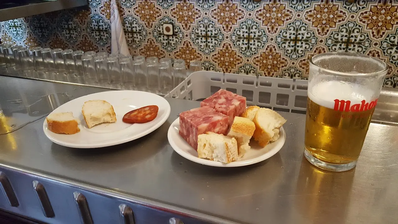 Bar counter with plates of chorizo slice, cubed cured sausage and bread, next to a glass of Mahou beer