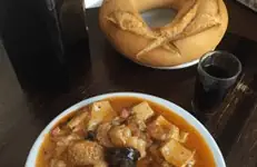 Bowl of stew with chunks of fish or meat, potato and olives in tomato sauce, next to a round loaf of bread