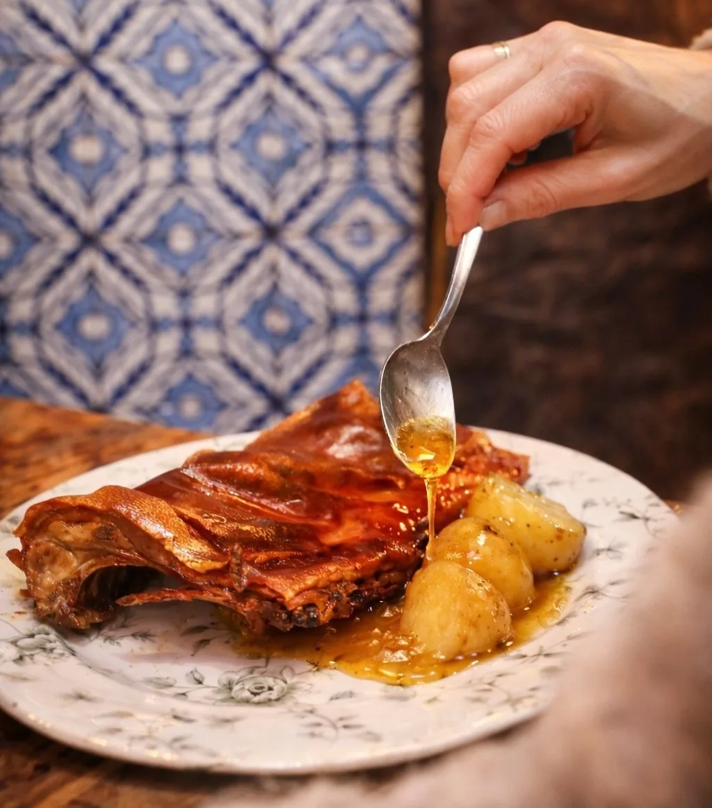 A spoon drizzling sauce over a portion of roast meat with crackling skin and potatoes on a floral plate