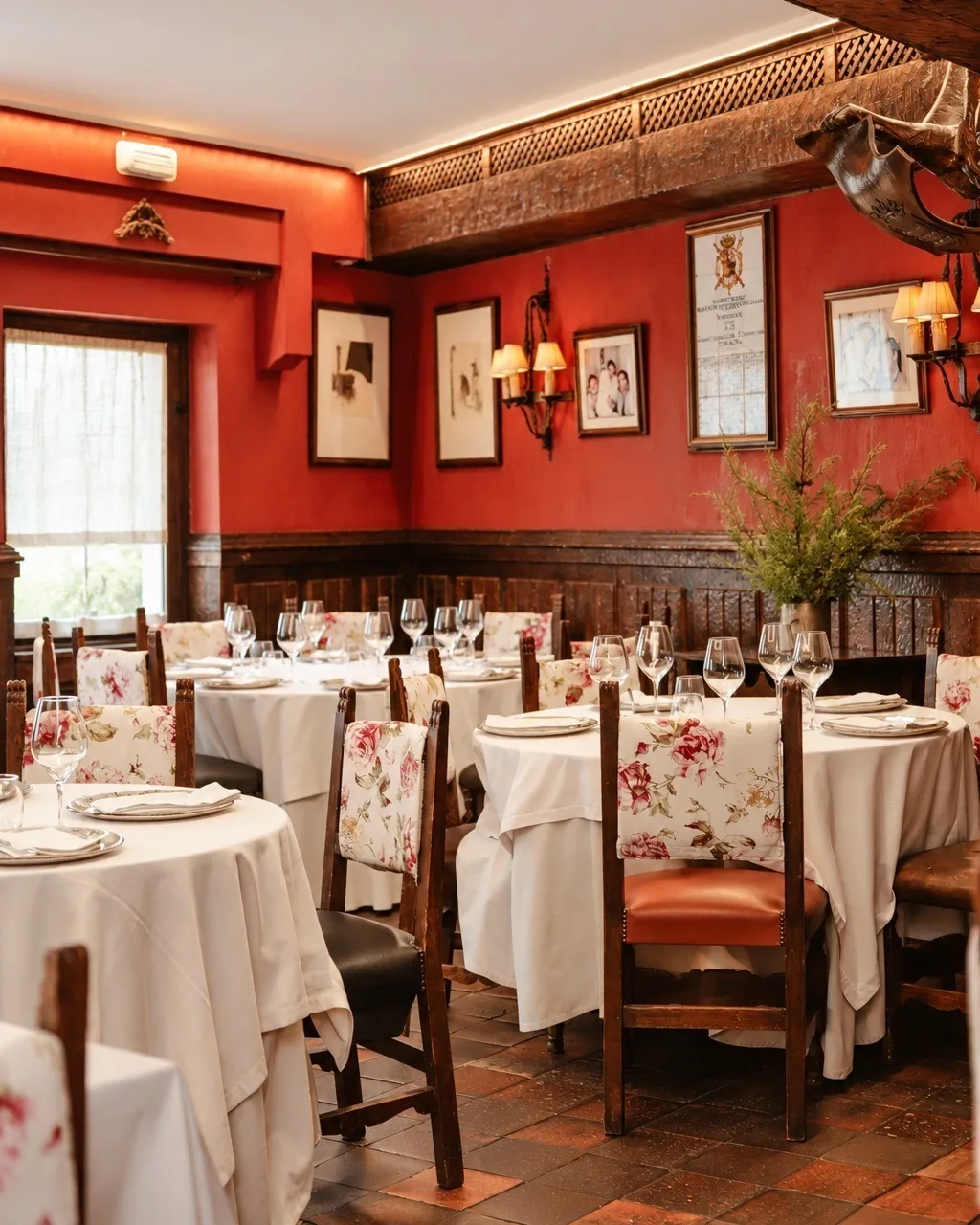A dining room with red walls, dark wood panelling, round tables with white cloths and floral chairs.