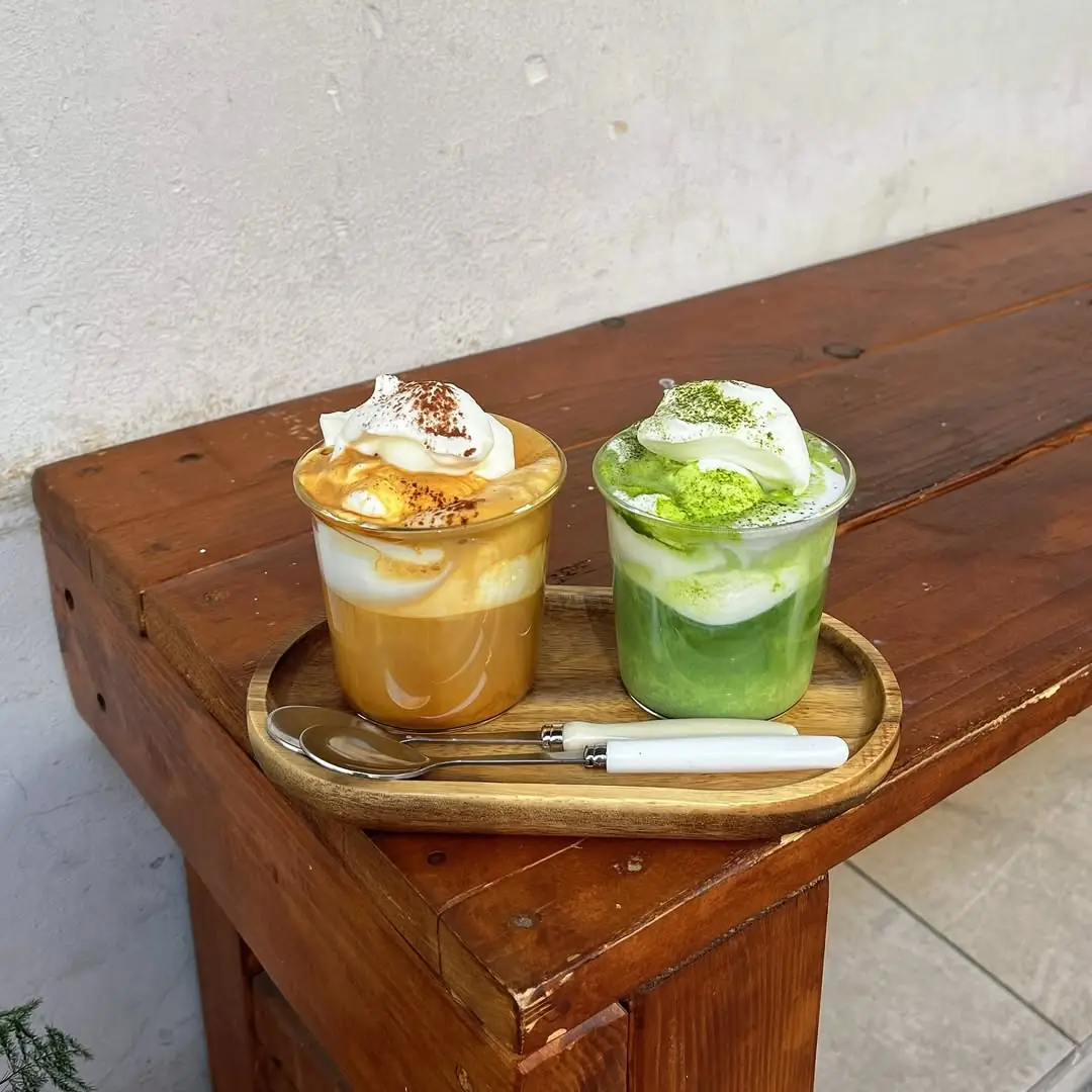 Two iced dessert drinks, one caramel-colored and one matcha, topped with whipped cream on a wooden tray