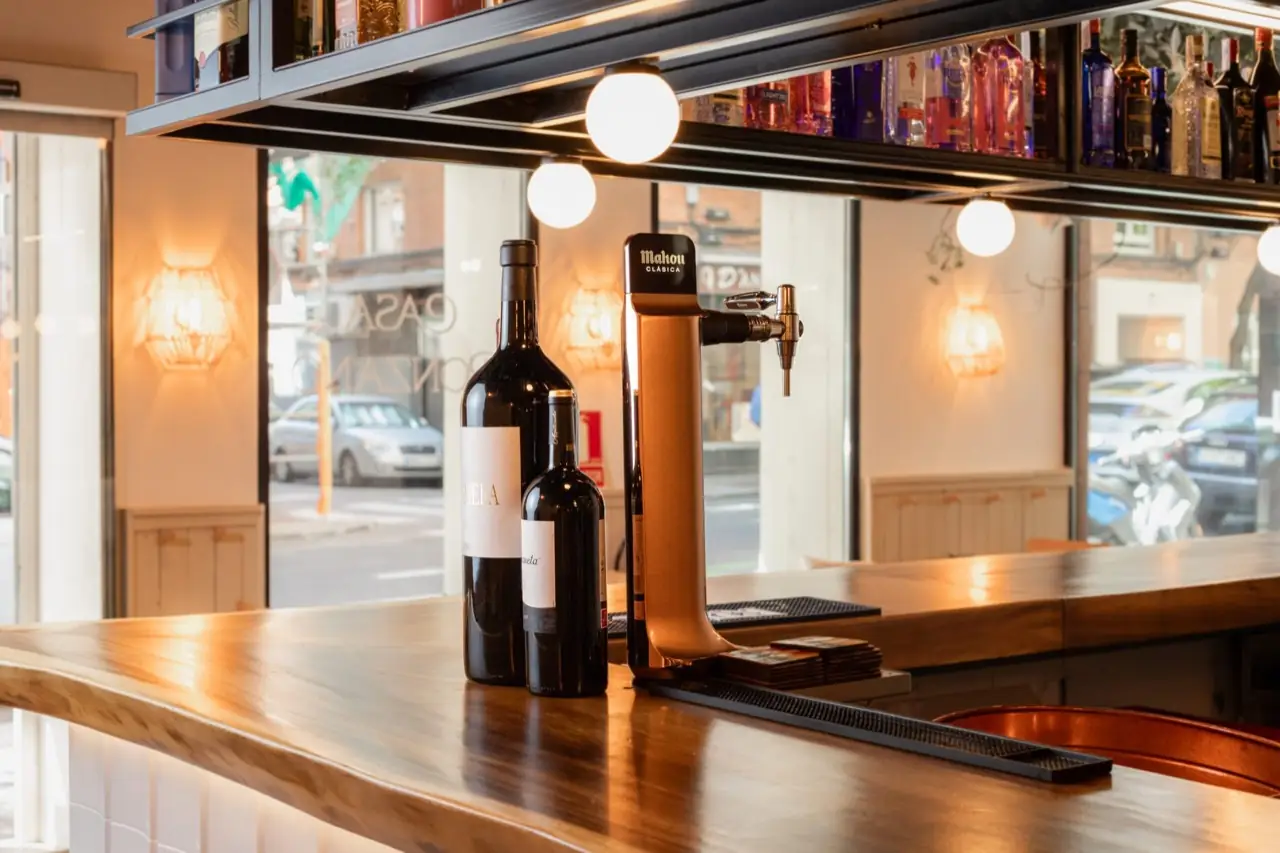 Wooden bar counter with two wine bottles and a copper Mahou beer tap, back-bar spirit shelves and a street-facing window