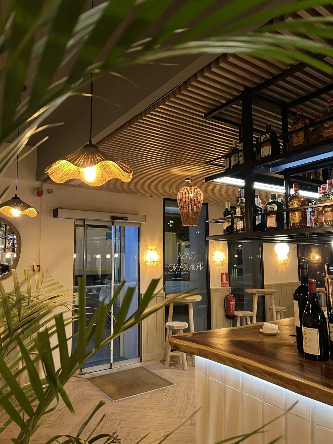 Casa Ponzano interior with woven pendant lamps, a wooden bar with wine bottles and spirit shelves seen past a palm