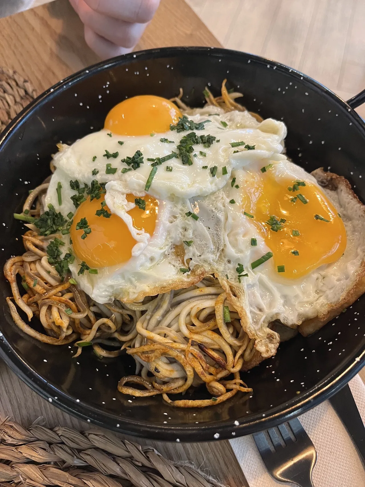 Three fried eggs with runny yolks sprinkled with chives over fried strands in a black speckled pan