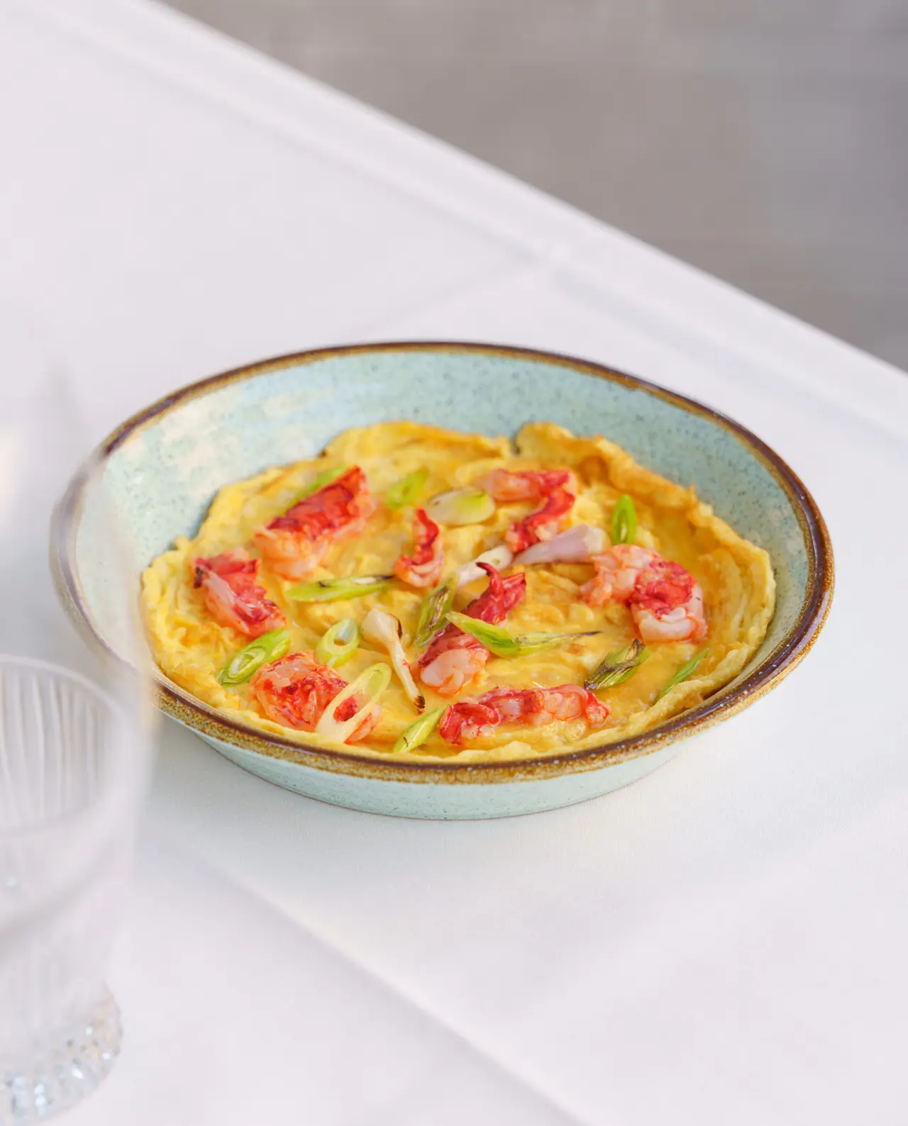 Open omelette studded with prawns and charred spring onions in a pale green-rimmed ceramic dish on a white tablecloth.