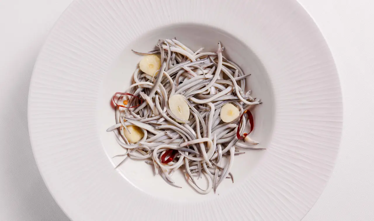 Nest of tiny elver-style baby eels with sliced garlic and dried chilli rings in a white bowl.