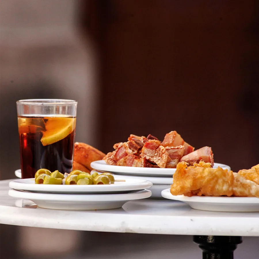 A round marble pedestal table set with a glass of dark vermut over ice with an orange slice, a small plate of pitted green olives on a cocktail stick, a plate of crisp-edged torreznos and a plate of golden battered fritters