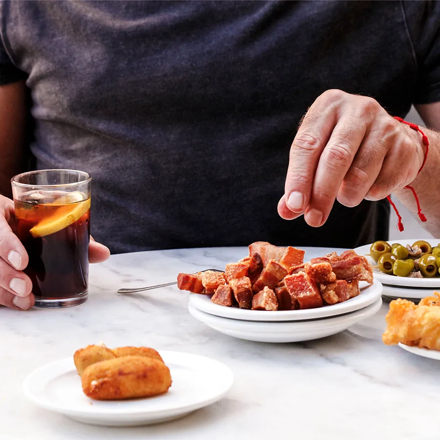 A marble table from the diner's own view: one hand holding a glass of dark vermut with an orange slice, the other reaching over a plate of cubed torreznos, with a plate of green olives and a plate of croquettes alongside