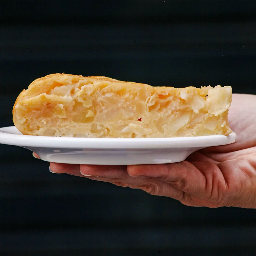 A hand holding out a small white plate carrying a thick wedge of potato tortilla, its centre soft and barely set, against a dark background