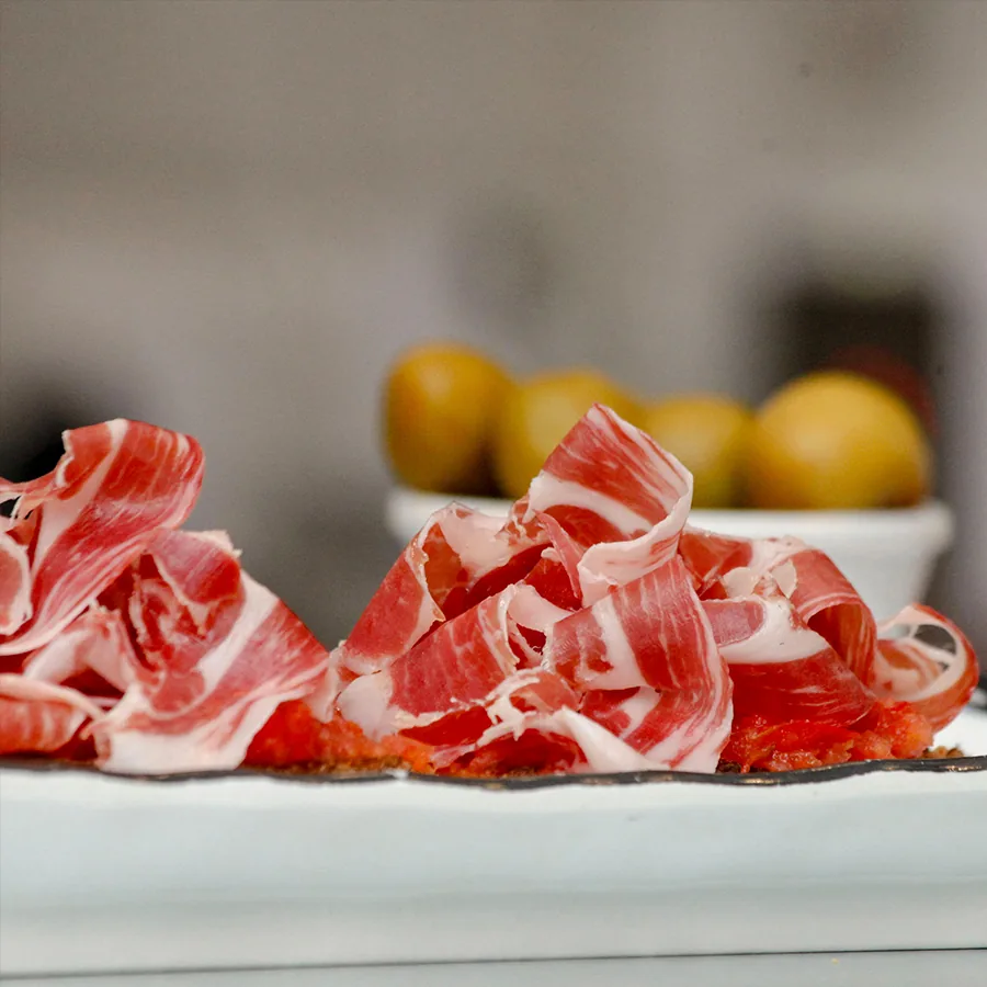 Ruffled slices of marbled cured ham laid over tomato-rubbed toast on a white plate, with a bowl of green olives blurred behind