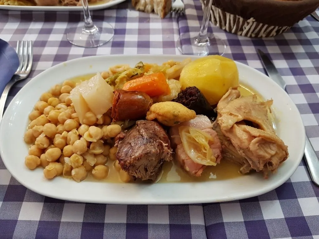 A white oval plate of chickpeas, potato, carrot, cabbage, chorizo, black pudding and boiled meats on a blue checked tablecloth