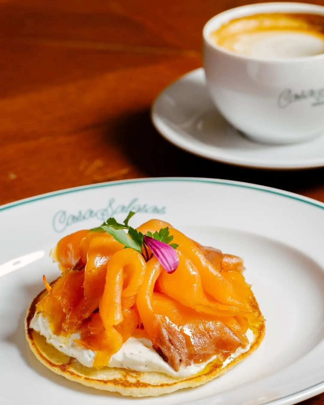 A small pancake topped with smoked salmon and a small flower, on a plate beside a cup of coffee