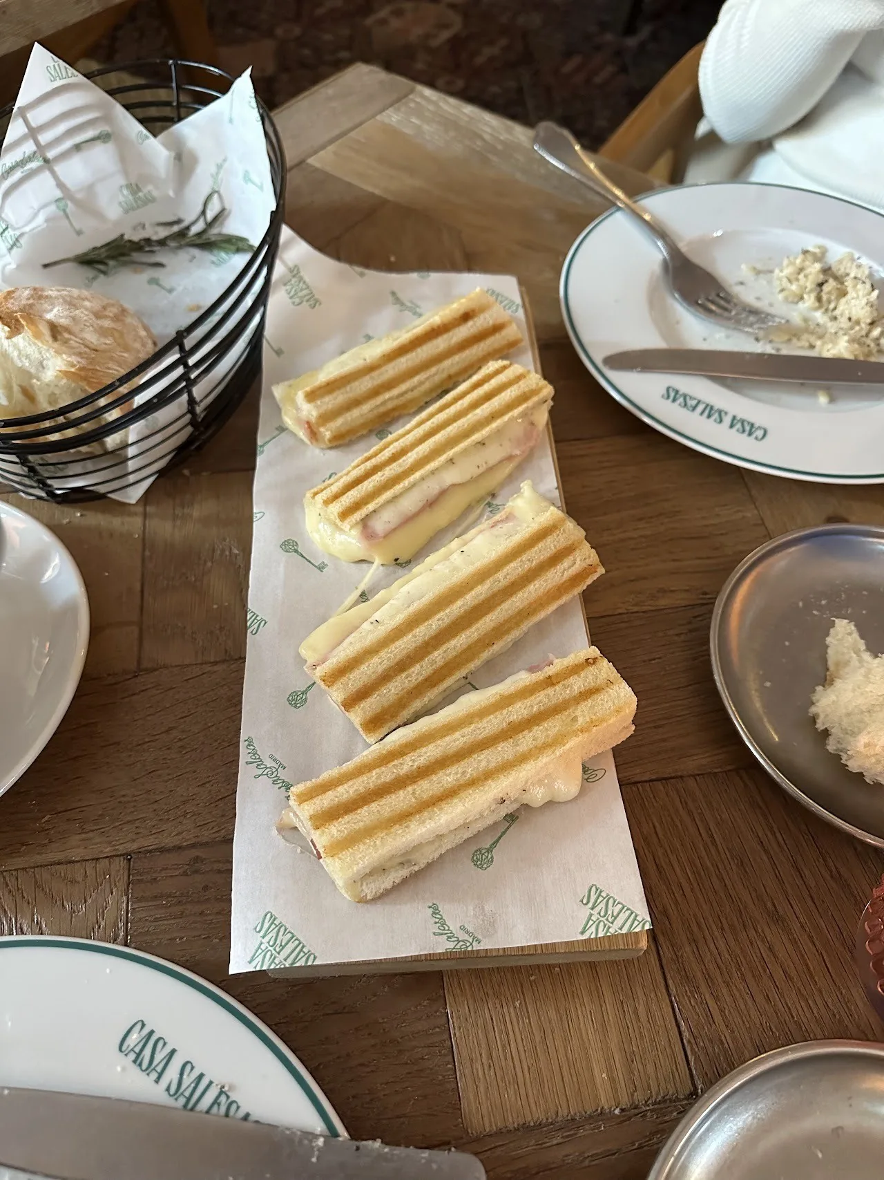 Four toasted ham and cheese sandwich strips on a paper-lined board beside a basket of bread
