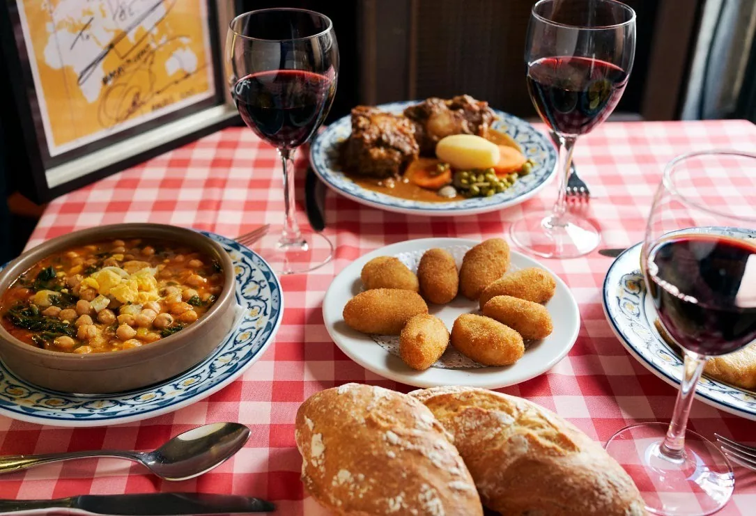 A red-and-white checked table with a stew pot, plates of croquetas and a stew dish, and glasses of red wine