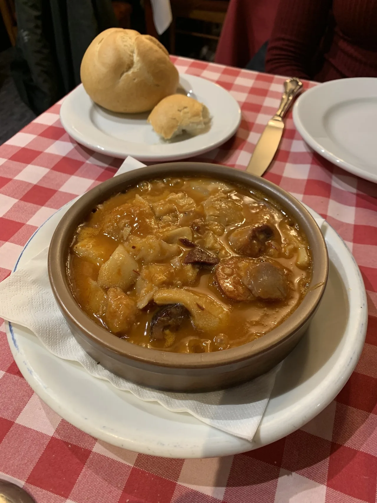 A brown earthenware dish of tripe stew with chorizo and black pudding on a red gingham tablecloth, with bread rolls behind