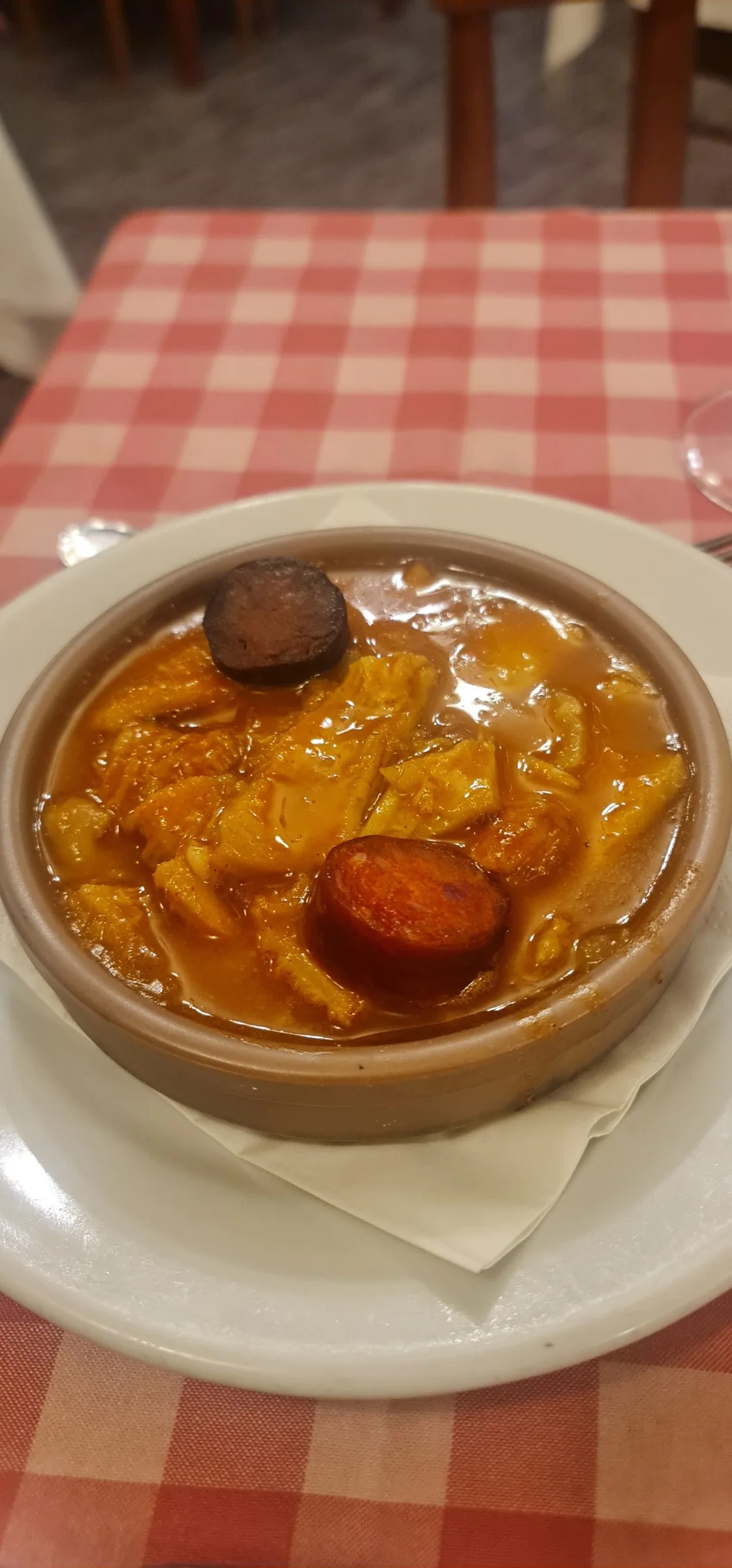 A small earthenware dish of tripe stew with a slice of black pudding and a slice of chorizo on a gingham tablecloth