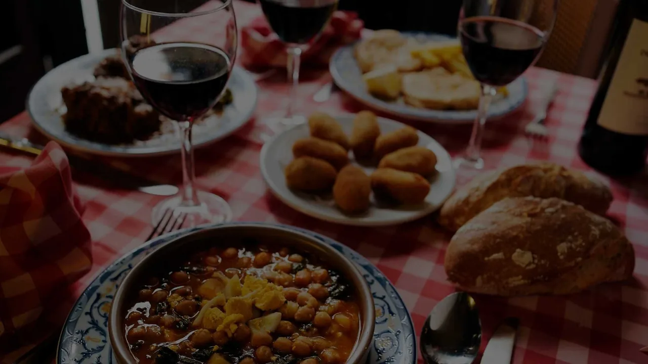 A table with a stew in an earthenware dish, croquettes and glasses of red wine on a checked cloth