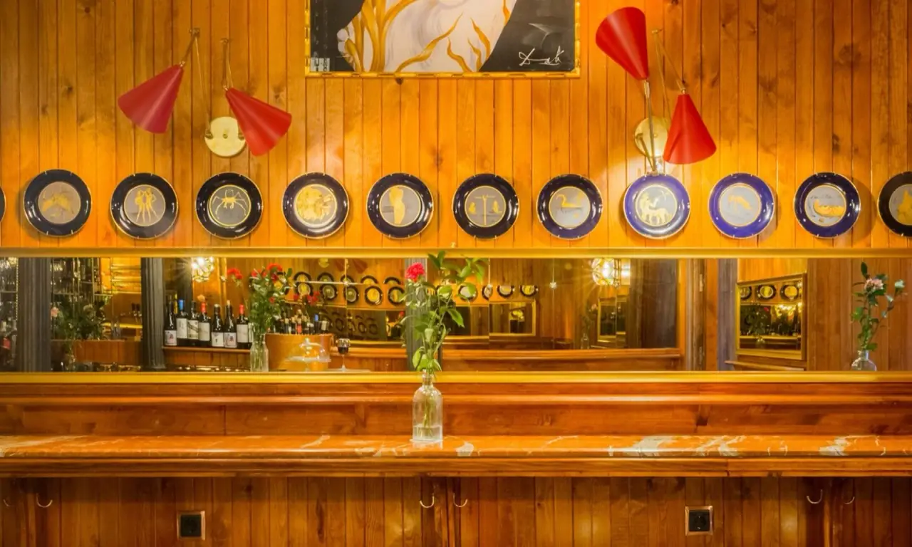 Wood-panelled bar interior with a row of blue-and-gold zodiac plates, red cone wall lamps and a mirrored back bar lined with wine bottles
