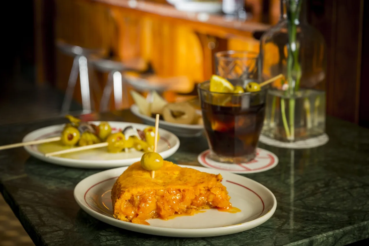 A wedge of potato omelette on a small plate beside olives and a glass of dark drink