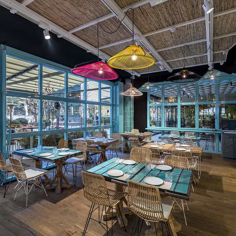 Restaurant dining area with teal tiled tables, rattan chairs, bamboo ceiling and colorful woven pendant lamps in pink and yellow