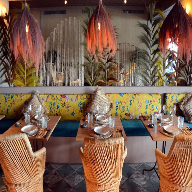 Restaurant booth seating with yellow floral upholstery, woven pendant lights and rattan chairs, tables set with glassware against tropical foliage backdrop