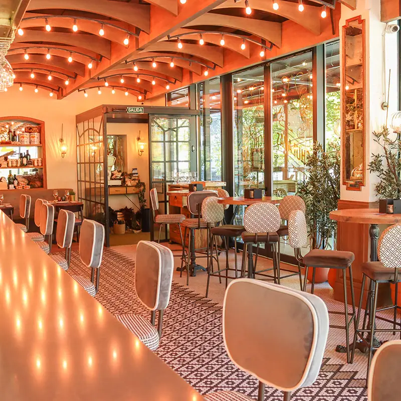 Restaurant bar and seating area with string lights along curved wooden ceiling beams, patterned upholstered stools and glass storefront