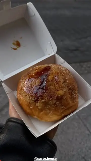 Sugar-dusted round brioche bun glazed with jam, held in an open white takeaway box over a grey pavement
