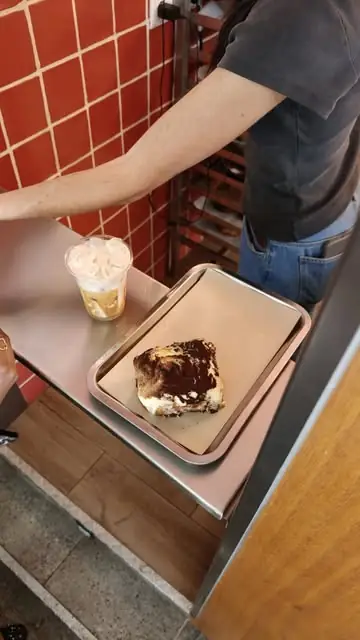 Slice of tiramisu dusted with cocoa on a pink metal tray beside an iced coffee, against a terracotta-tiled counter