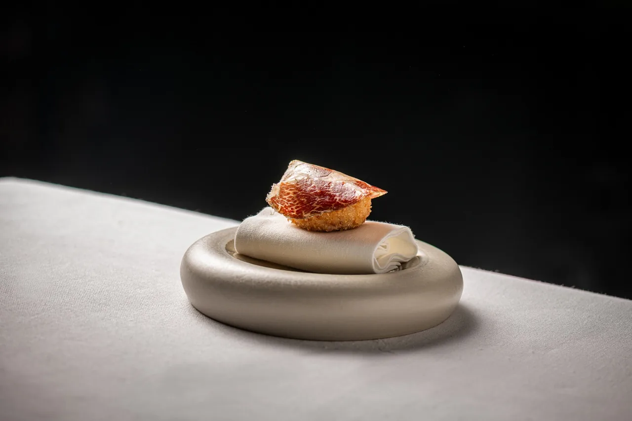 A small round croquette-like bite on a curved cream dish against a dark background