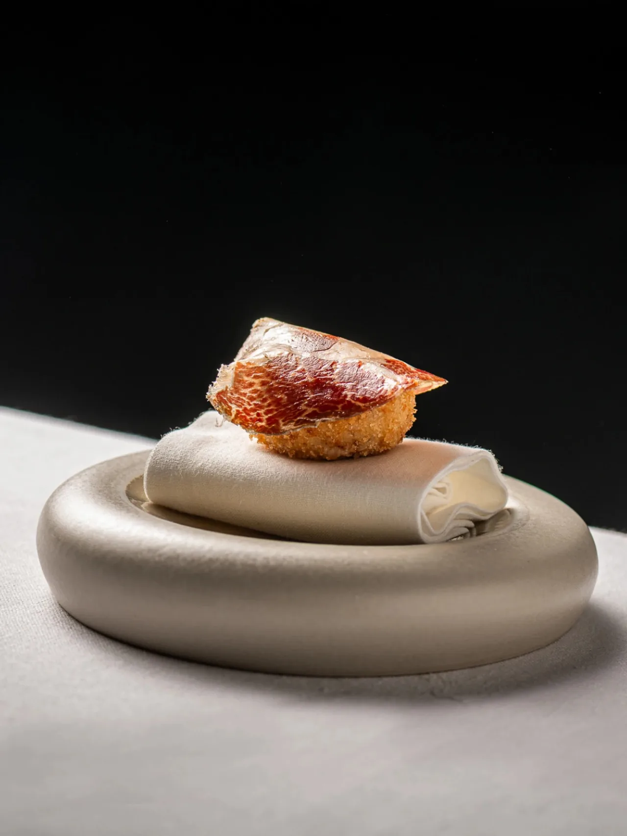 A curled cream-coloured plate holding a small round fried bite with a red top on a white surface