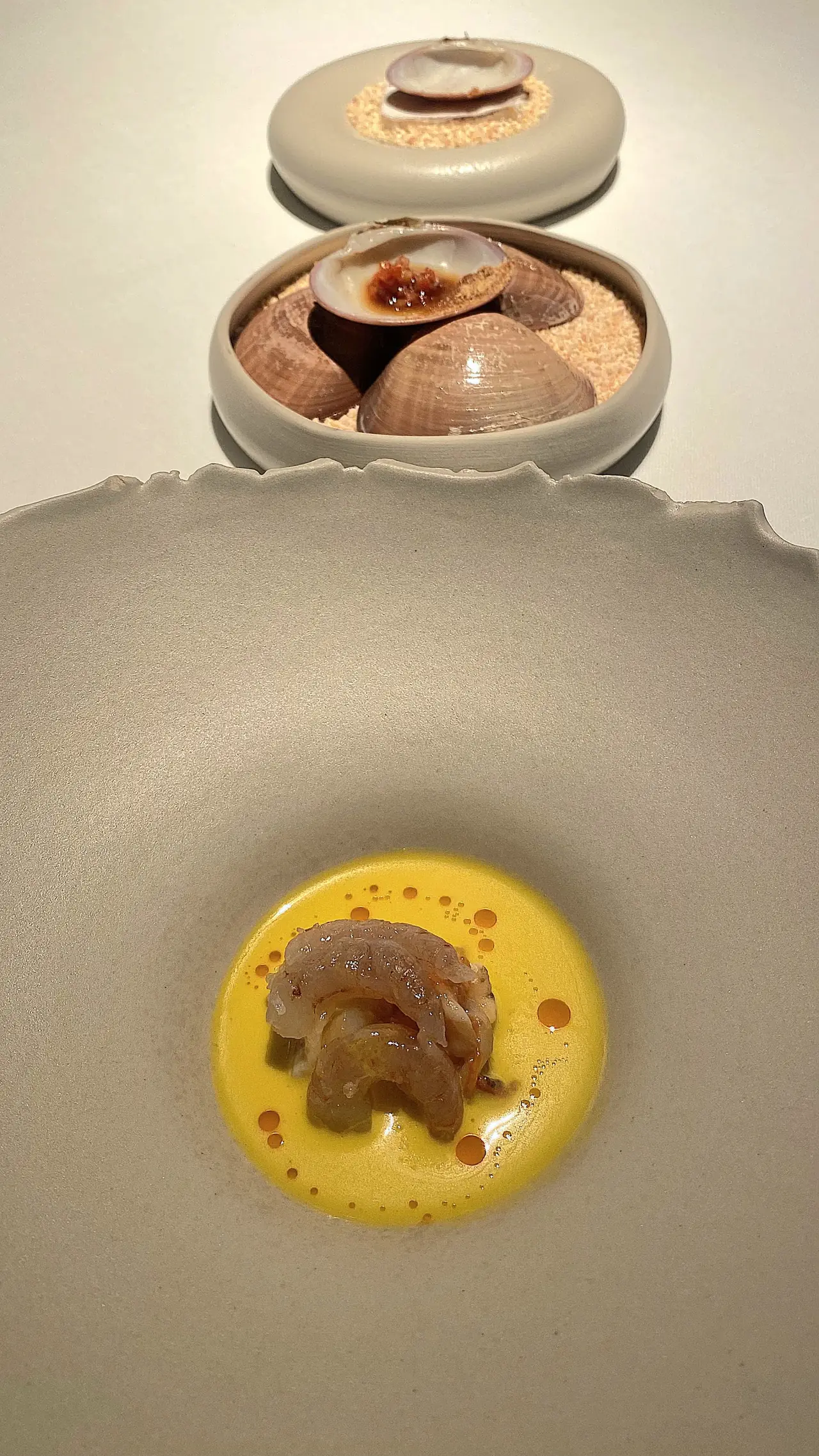 Prawn on yellow sauce in a bowl, with a dish of clams in the background