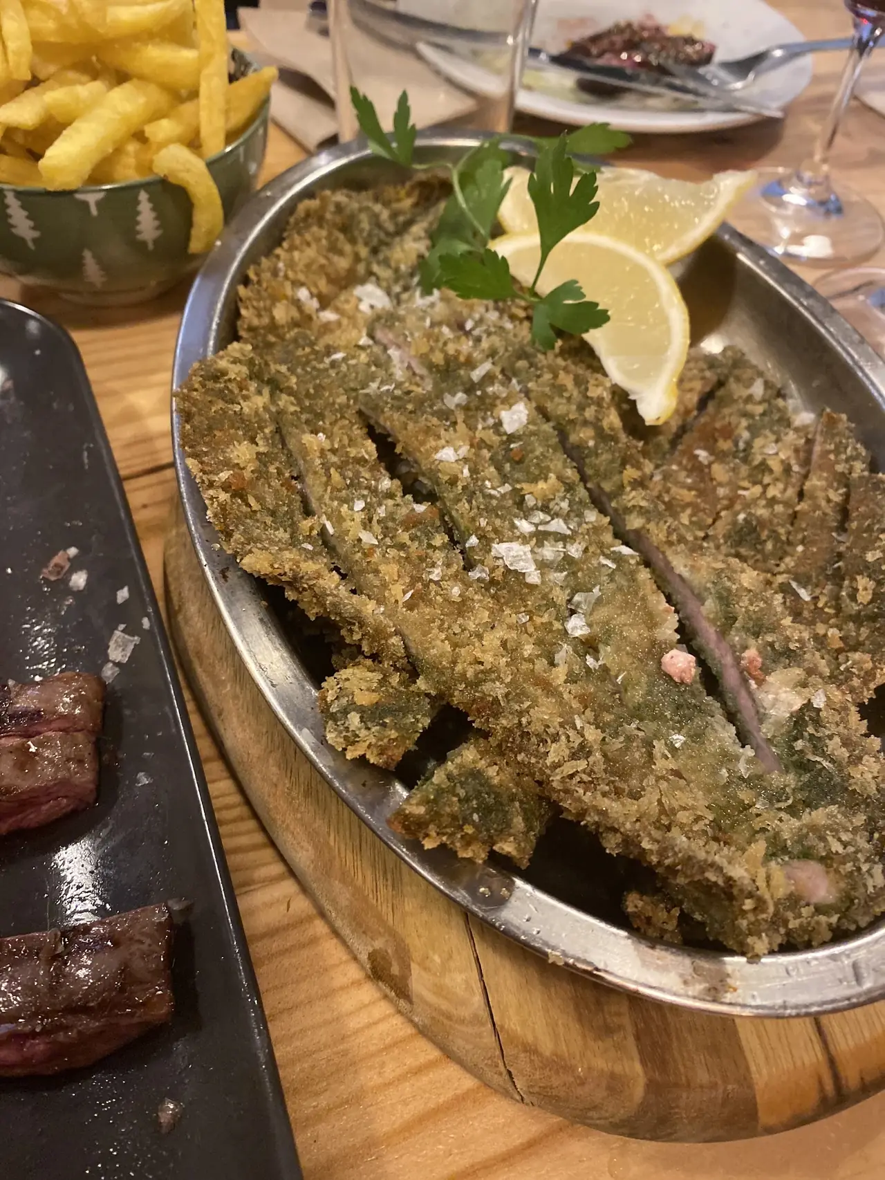 Breaded fried anchovy fillets with sea salt and lemon wedges in a metal dish, steak in background