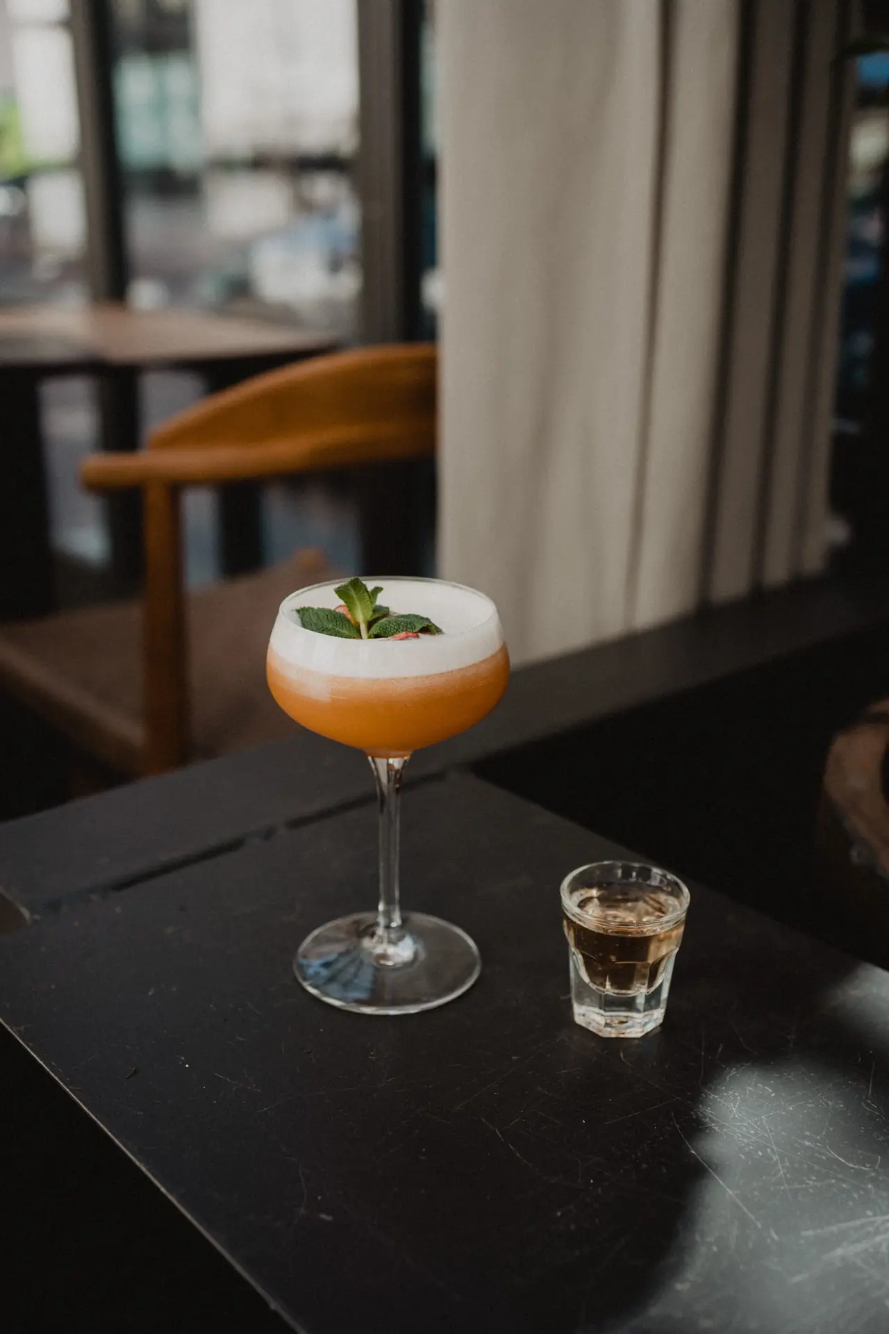 Orange cocktail with white foam top garnished with mint, next to a shot glass of amber liquid