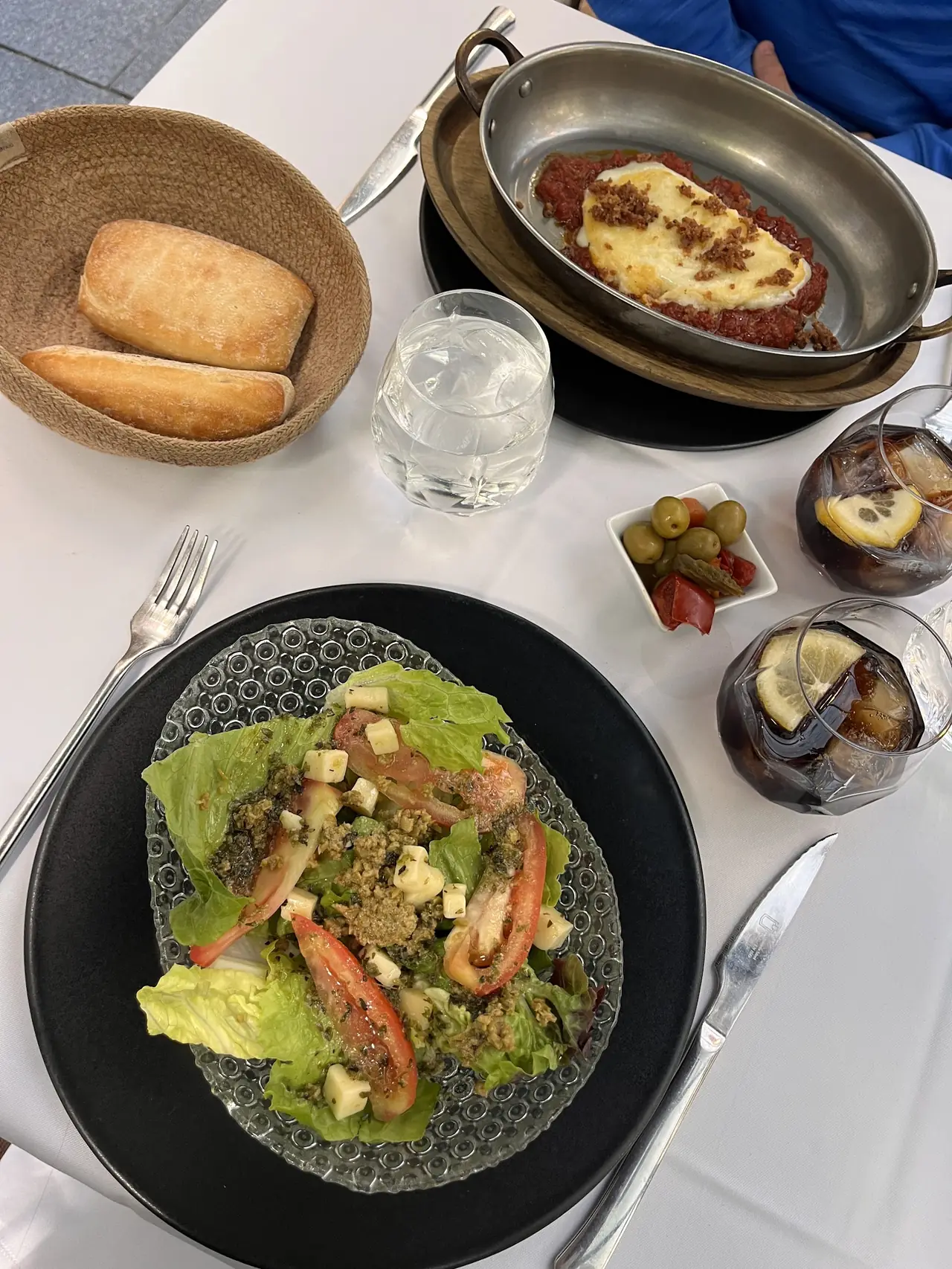Table spread with tomato salad, baked cheese in tomato sauce, bread basket, olives and cuba libre cocktails