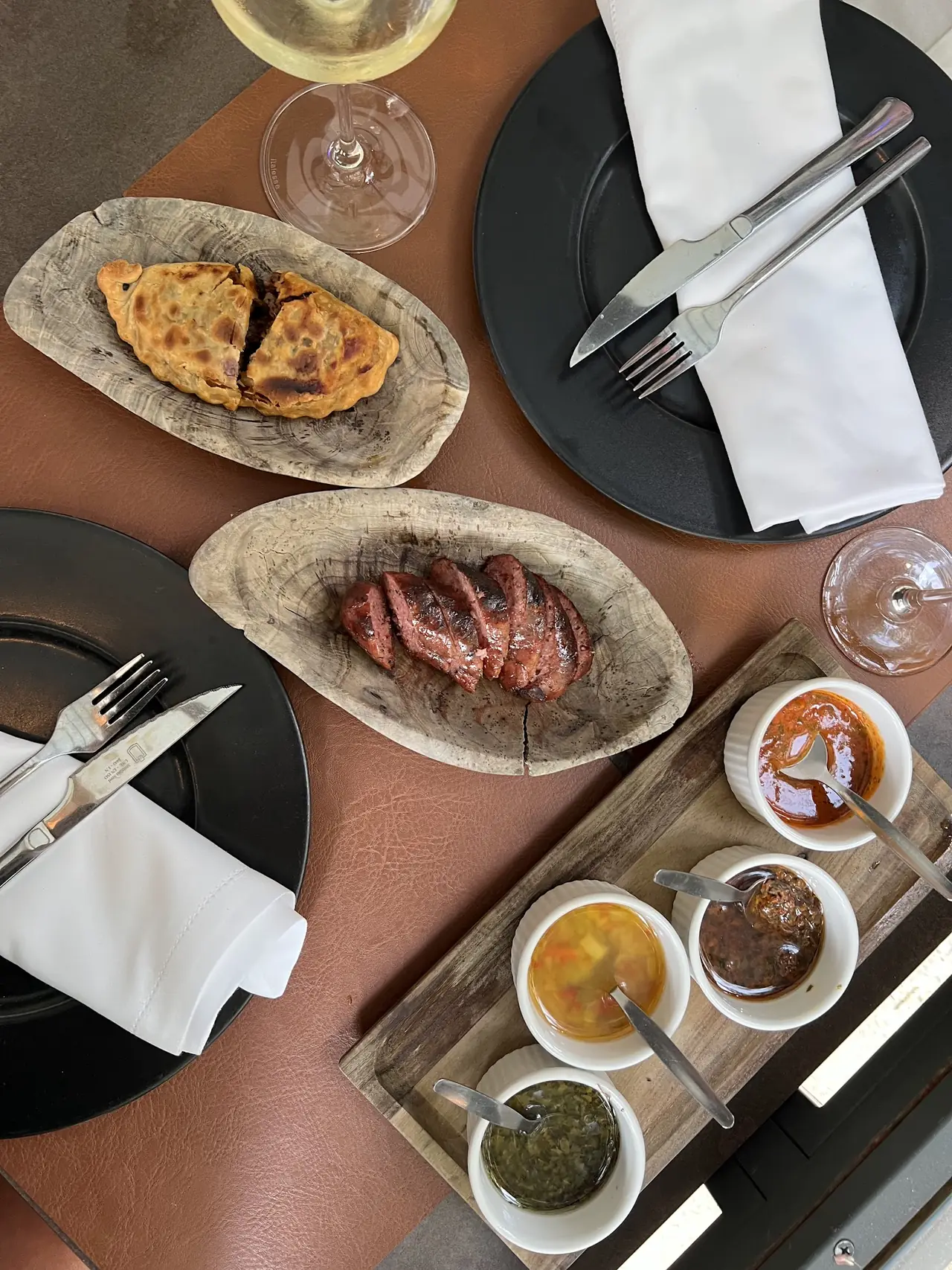 Overhead view of grilled sausage and empanada in wooden dishes, with three dipping sauces on a board